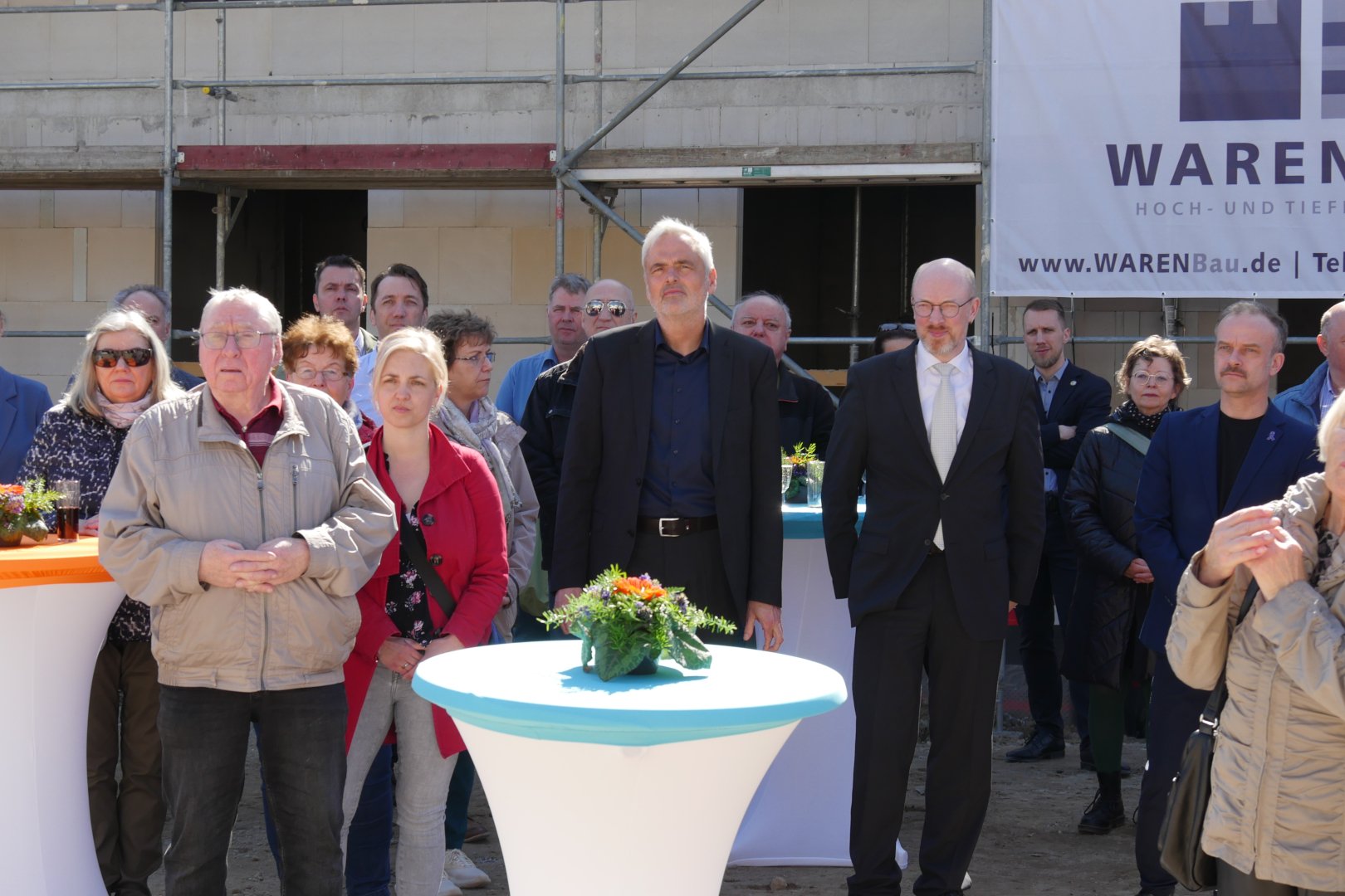 Personen hören einem Redner zu, sie stehen verteilt zwischen Tischen
